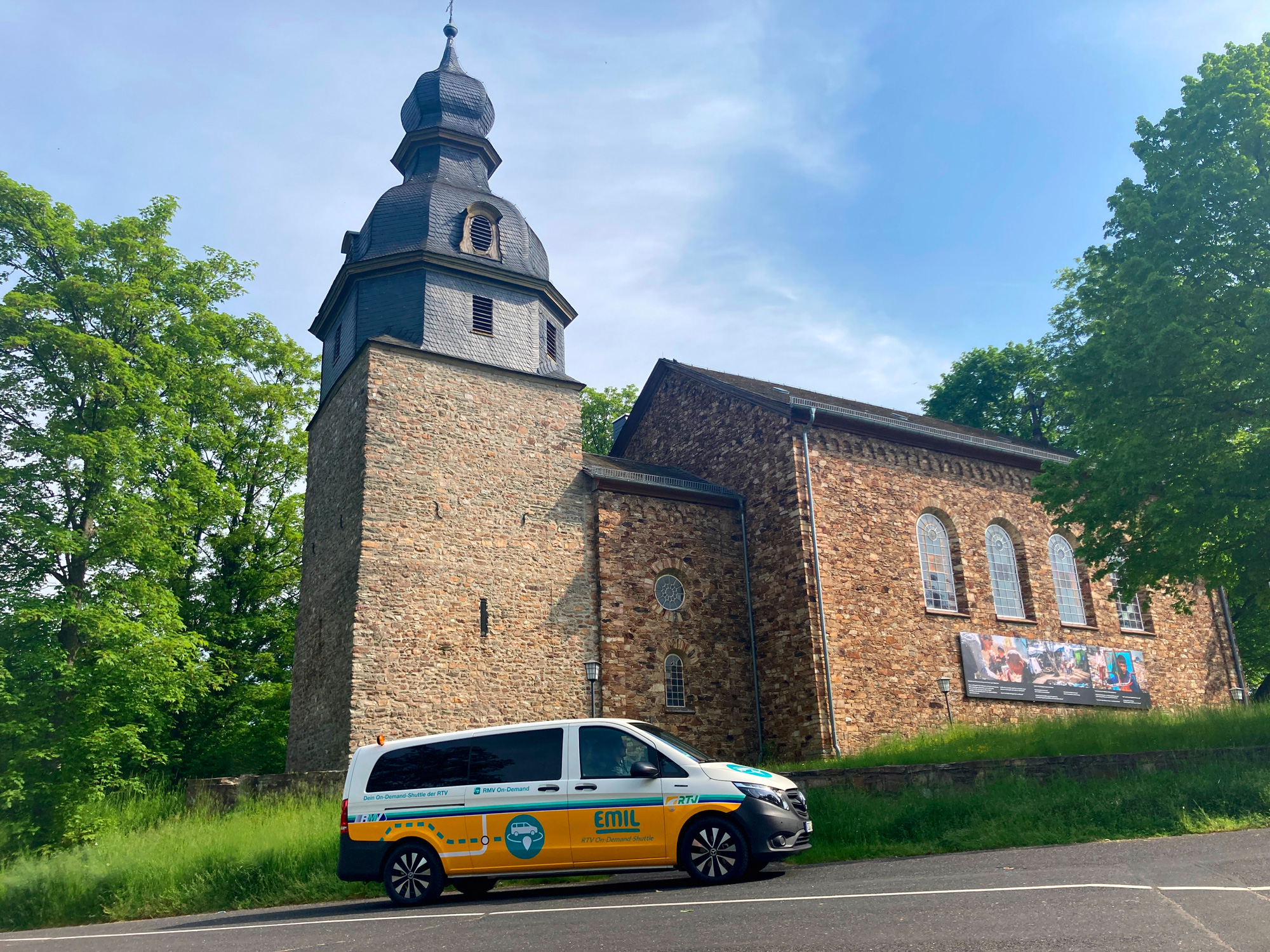 Gelb-weißes EMIL-Shuttle der Rheingau-Taunus-Verkehrsgesellschaft geparkt vor einer alten Bruchsteinkirche mit Schieferdach und Rundbogenfenstern, eingebettet in grüne Bäume.